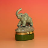 Side view of ceramic elephant trinket box featuring a trunk-up elephant standing on a rocky base with gold clasp.