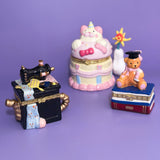 Group photo of three hand-painted ceramic trinket boxes — a sewing machine, graduation bear, and pastel cat cake — displayed on a purple background.